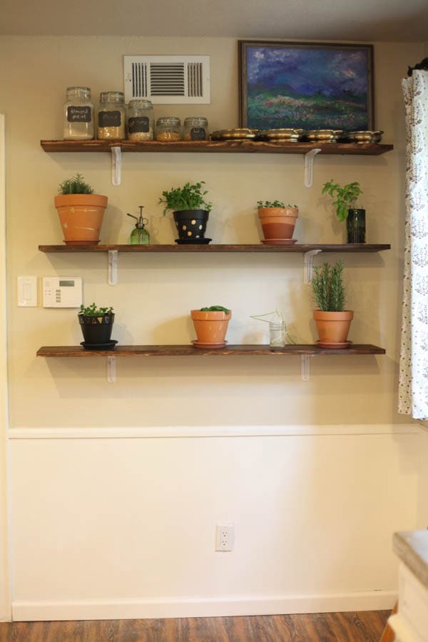A DIY herb wall with potted fresh herbs and a landscape style painting on wooden shelves. 