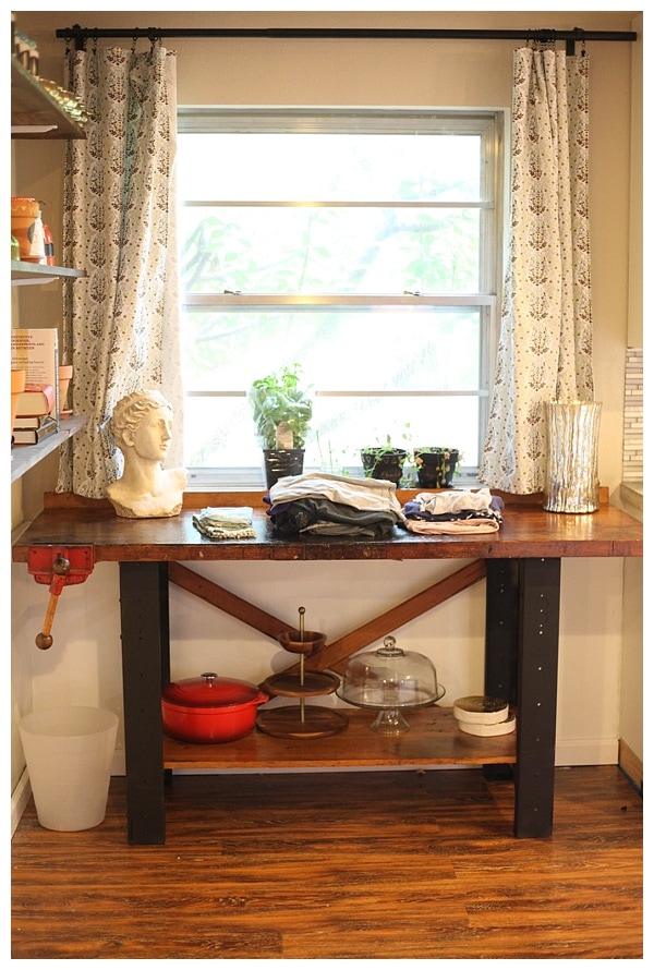 A wooden table in a clean kitchen, with folded clothes on top. 