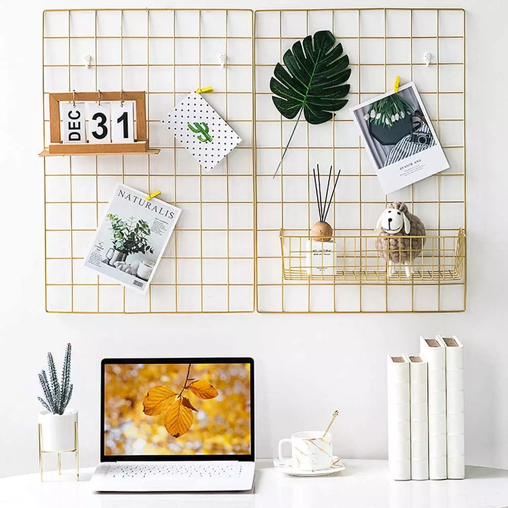 A gold organizational wall grid covered in small art prints and a calendar, above a white desk topped with a laptop, small plant, cup of tea and decorative books. 