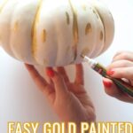 Hand holding a white pumpkin and using a gold leaf pen to add polka dots.