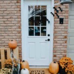 Front door decorated for halloween. White door with a black wreath, bats and spider webs. Orange and white pumpkins along with orange and yellow colored mums on a black and white striped doormat and another doormat layered that reads witch please.