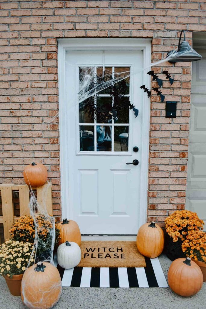 Front door decorated for halloween. White door with a black wreath, bats and spider webs. Orange and white pumpkins along with orange and yellow colored mums on a black and white striped doormat and another doormat layered that reads witch please. 