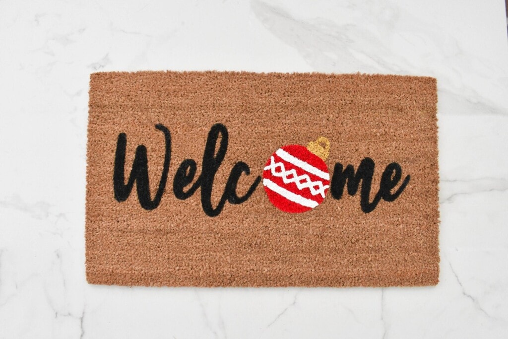 a doormat that says "welcome" with a Christmas ornament in place of the O