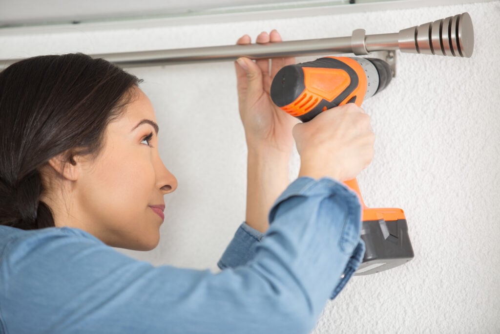 Woman with a drill hanging a curtain rod.