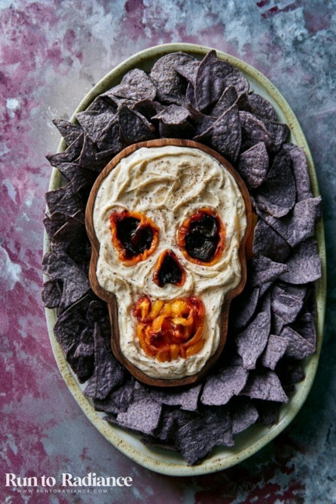 Skull-shaped Halloween charcuterie board dip with roasted pepper eyes and blue corn tortilla chips